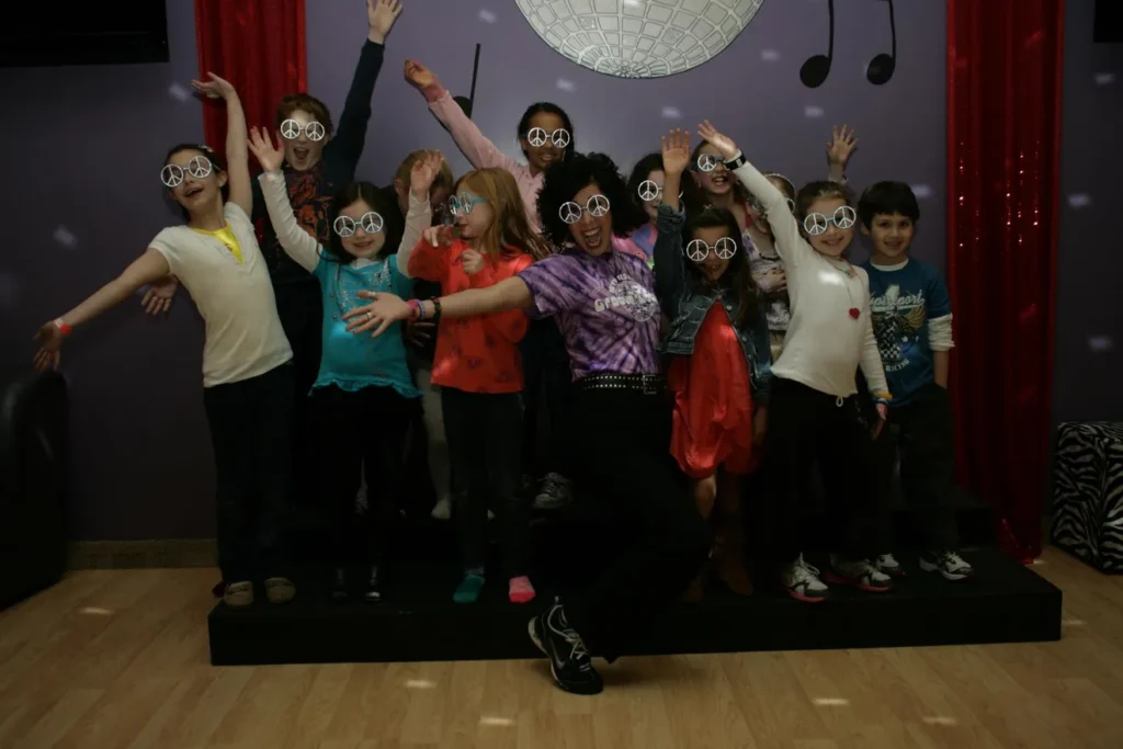 A group of children and adults joyfully posing indoors with raised arms.