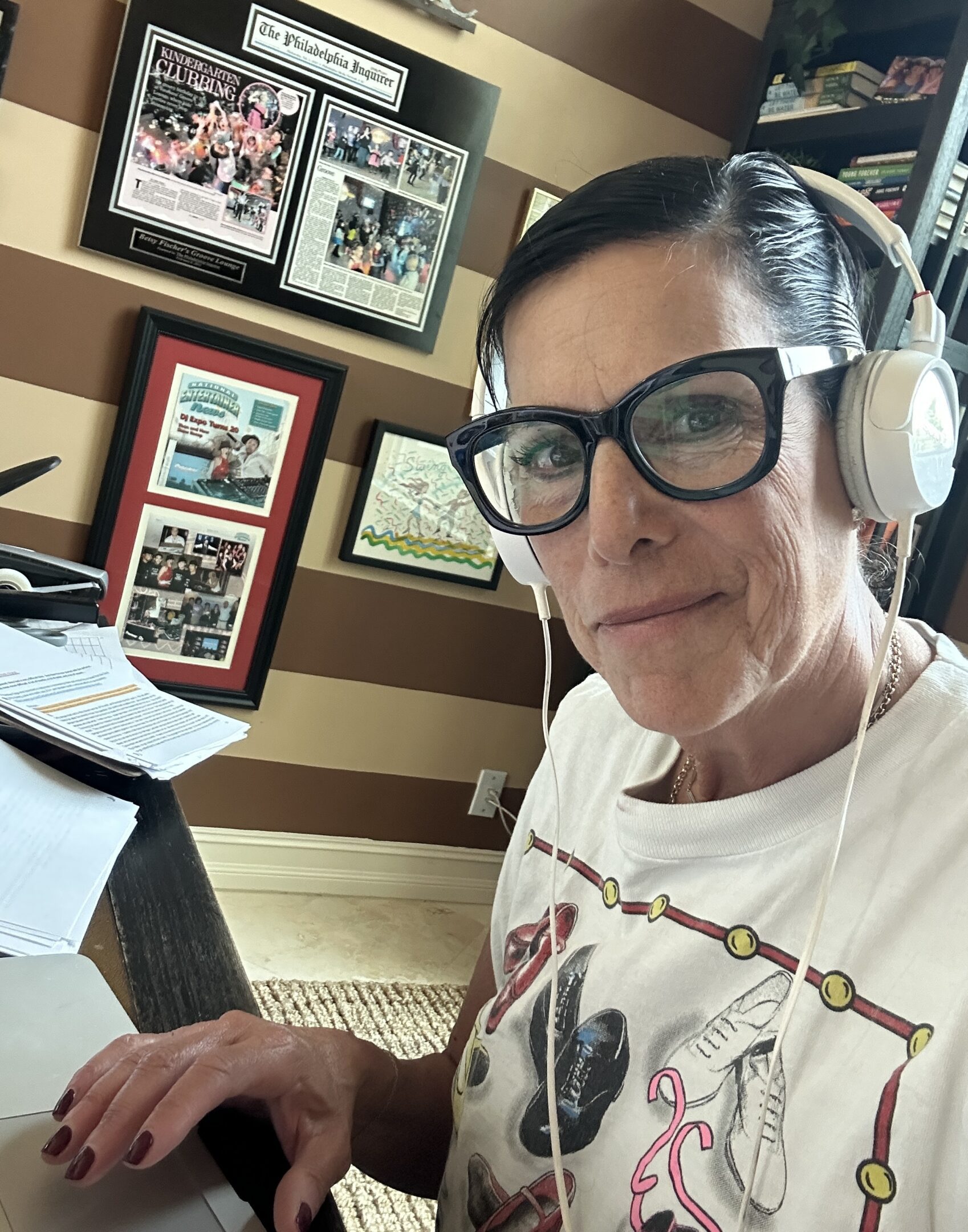 Person wearing glasses and headphones, sitting at a desk with framed pictures on the wall.