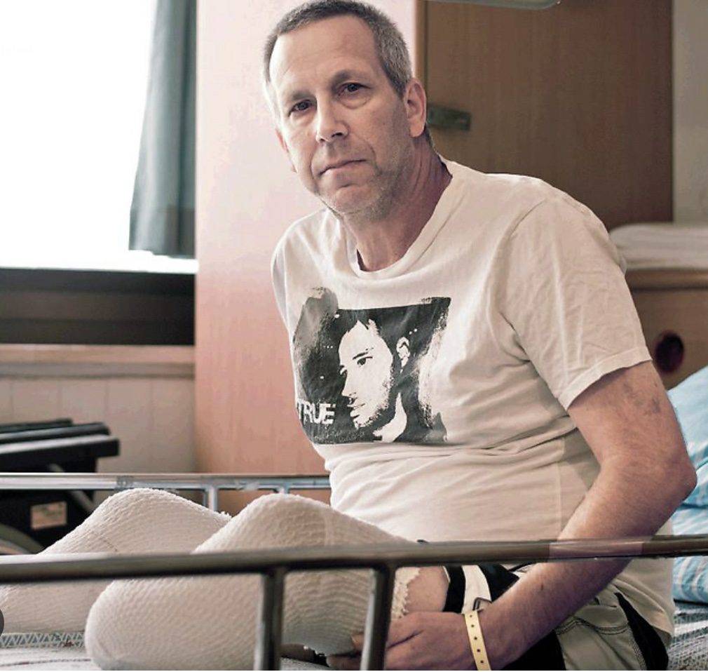 Man sitting on a bed in a casual room, wearing a printed t-shirt.