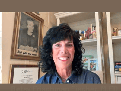 Smiling woman with curly black hair in a cozy room with books and framed art.