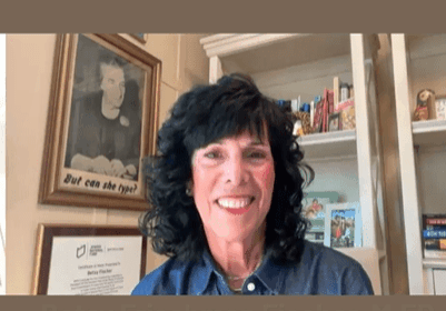 Smiling woman with curly black hair in a cozy room with books and framed art.